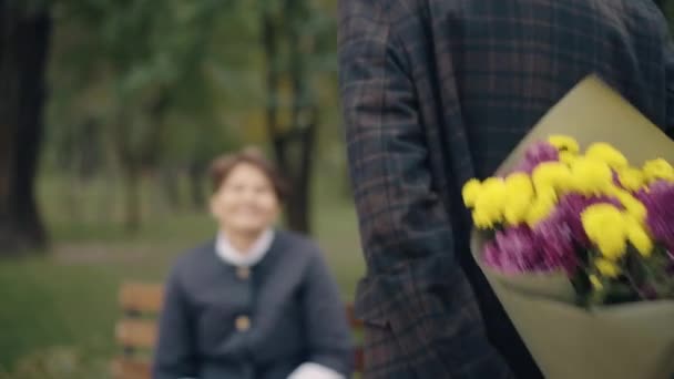 ラックフォーカスは、ギフトを受け取る幸せな女性の顔にシニア男性の手の花の花束から行く。愛する白人夫驚くべき妻日付秋や春の公園屋外で. — ストック動画