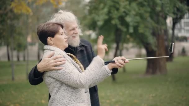 Sidovy över moderna par pensionärer med videochatt på smartphone utomhus. Positivt kramande vit man och hustru viftar i telefon och pratar. Koncept för trådlös kommunikation. — Stockvideo