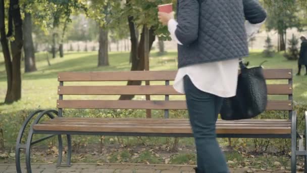 Beztroska starsza kobieta siedząca na ławce w Sunny Park pijąca kawę na wynos. Portret zrelaksowanego, pięknego, białego emeryta odpoczywającego na świeżym powietrzu jesienią. Koncepcja stylu życia i wypoczynku. — Wideo stockowe