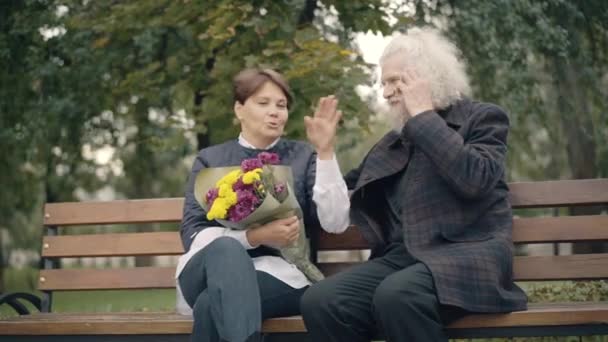 Pozytywna zrelaksowana para białych emerytów siedząca na ławce w jesiennym parku rozmawiająca. Szczęśliwy beztroski mężczyzna i kobieta cieszący się szczęśliwy wypoczynek na świeżym powietrzu. Pojęcie miłości i randkowania. — Wideo stockowe