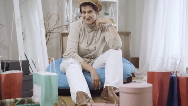 Die Kamera nähert sich der glücklichen selbstbewussten Kaukasierin mittleren Alters, die auf einem Taschensofa mit Einkaufstaschen auf dem Boden sitzt. Porträt einer eleganten, schönen Shopperin, die zu Hause posiert. — Stockvideo