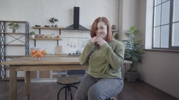 Elbűvölő, félénk, vörös hajú, kaukázusi nő ül a széken a konyhában, és a kamerát nézi. Egy gyönyörű pufók hölgy portréja, aki otthon pózol. Életmód-koncepció. — Stock videók