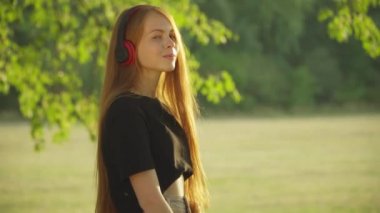 Güneş ışığının altında dikilmiş kameraya gülümseyen, kulaklıklı, beyaz bir kızın yan görüntüsü. Yaz parkında güneş ışığında poz veren güzel bir gencin portresi..