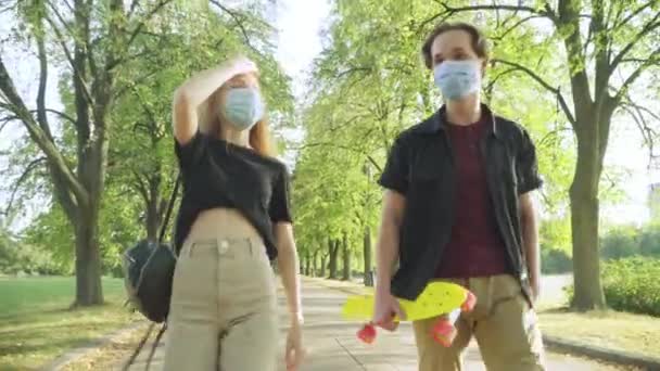 Cheerful relaxed Caucasian teenagers walking in sunny summer park in Covid-19 face masks talking. Portrait of joyful carefree teen boy and girl resting outdoors on coronavirus pandemic lockdown. — Stock Video