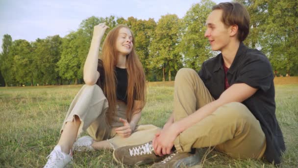 Pozitív nyugodt tinédzser pár ül a nyári mezőn beszélgetni és nevetni. Portré boldog gondtalan kaukázusi tinédzser fiú és lány társkereső szabadban naplementekor beszél és mosolyog. — Stock videók