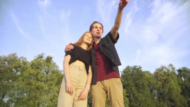 Glücklich liebender Teenager-Freund umarmt Freundin und zeigt weg, während er redet. Porträt einer entspannten kaukasischen Jugendlichen Paar Dating im Freien an einem sonnigen Tag mit schönem Himmel im Hintergrund. — Stockvideo