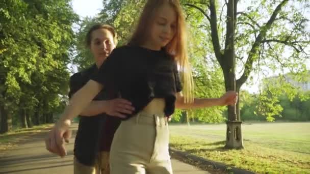 Sonriente adolescente asegurando chica patinaje a caballo en soleado parque al aire libre. Felices amigos caucásicos relajados divirtiéndose patinando a la luz del sol en el día de verano. Relajación y amistad. — Vídeos de Stock