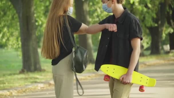 Menino e menina caucasiano adolescente irreconhecível em máscaras faciais Covid tocando cotovelos reunião no parque de verão. Adolescentes despreocupados relaxados com skate e mochila cumprimentando ao ar livre na pandemia de coronavírus. — Vídeo de Stock