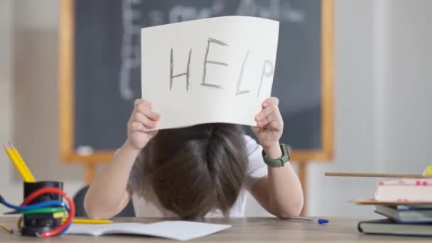 Utmattad söt skolpojke sätta huvudet på skrivbordet och höja Hjälp skriven på papper. Porträtt av trött uttråkad vit pojke sitter i klassrummet i skolan. Begreppet utmattning och trötthet. — Stockvideo