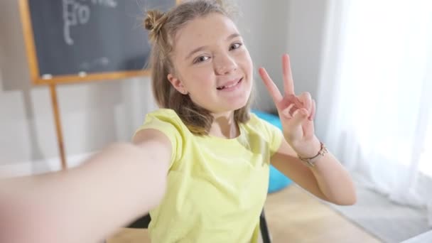 Vrolijk mooi blank meisje neemt selfie in de klas kijken naar de camera en tonen vrede gebaar. Portret van een charmant positief schoolmeisje dat plezier heeft in de pauze op school. Generatie Z. — Stockvideo