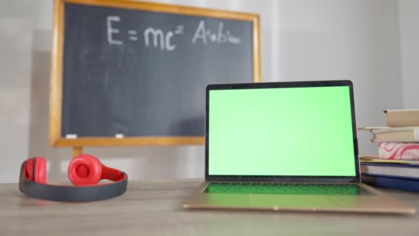 책상에 크롬 스크린이 있는 노트북을 클로즈업 한 뒤 헤드폰을 쓰고 칠판을 배경으로 흐릿하게 만들었다. 학교 실내의 녹색 스크린 장치입니다. 현대 과학 기술의 개념. — 비디오