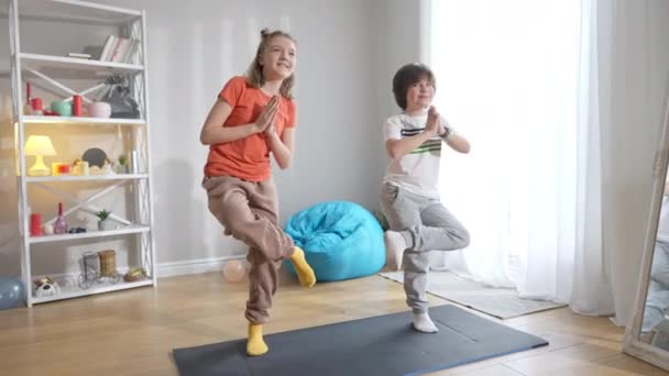 Weitwinkelporträt eines fröhlichen kaukasischen Jungen und Mädchens, das in Baumpose auf einer Trainingsmatte steht. Fröhliche Geschwister oder Freunde, die zu Hause Yoga-Übungen machen. Training des kleinen Yogi. — Stockvideo