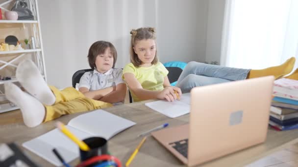 Szerokie ujęcie zrelaksowanych białych dzieci ze stopami na stole patrzących na laptopa i mówiących. Pozytywny beztroski uczeń i uczennica odrabiają lekcje w domu. Koncepcja zaufania i edukacji. — Wideo stockowe