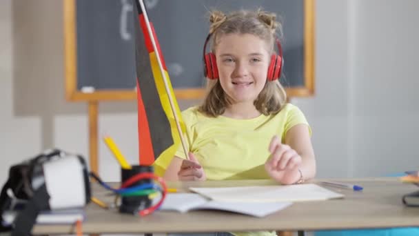 Retrato de encantadora colegial alemã sorridente em fones de ouvido sentados na mesa em sala de aula segurando bandeira nacional. Menina caucasiana confiante bonito gostando de educação na escola. Orgulho e patriotismo — Vídeo de Stock