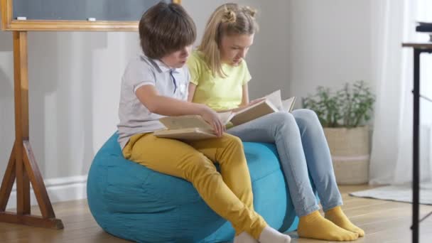 Ampla foto de menino e menina caucasianos sentados em cadeira confortável saco em livros de leitura de sala de aula. Colegas inteligentes absorvidos que gostam de literatura na escola moderna. Estilo de vida e educação. — Vídeo de Stock