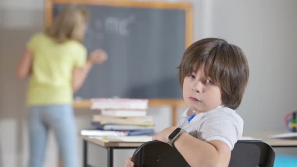 Stufo esausto studente caucasico girando a camera sospirando come scolaretta offuscata scrivere sulla lavagna sullo sfondo. Ragazzo bruna carino stanco di lezioni. Noia ed esaurimento. — Video Stock