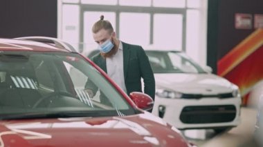 Covid yüz maskeli, düşünceli, başarılı, beyaz sakallı adam otomobil bayiliğinde otomobil seçiyor. Zengin, kendine güvenen işadamının portresi Coronavirüs salgınında araçları inceliyor..