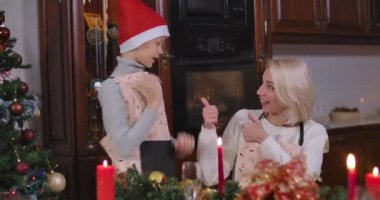 Önlüklü olumlu bir çocuk Noel şapkası takıyor, neşeli bir anne baş parmağını kaldırıyor ve gülümsüyor. Mutlu Kafkas ailesi yeni yıl kutlamalarına hazırlanıyor. Sinema 4k ProRes Merkezi..