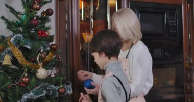 Kafkasyalı mutlu küçük oğlunun Noel ağacını evde annesiyle süslemesinin yan görüntüsü. Pozitif esmer çocuk ve genç sarışın kadın yeni yıl ağacına oyuncak asıyorlar. Sinema 4k ProRes Merkezi