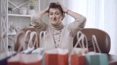 Orta yaşlı esmer kadın, el ele tutuşmuş, öndeki alışveriş çantalarına bakıyor. Kara Cuma 'nın satış gününde evde strese girmiş Kafkas alışveriş koliklerinin portresi.