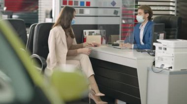 Covid-19 yüz maskeli güzel, zayıf bir kadın galerideki satıcıdan araba anahtarı alıyor. Heyecanlı Kafkasyalı başarılı iş kadınının portresi Salgın üzerine araba satın alıyor.