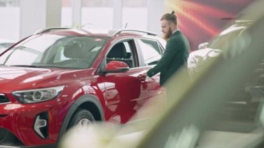 Kendine güvenen beyaz adamın galeride lüks kırmızı araba açması ve sürücü koltuğunda ağır çekimde oturması geniş açı. Varlıklı iş adamı galeride araç seçiyor..