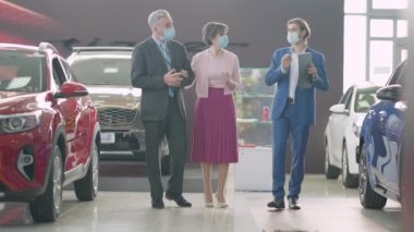 Müşterilerin ve Covid maskeli torbacıların geniş açılı görüntüsü galeride konuşuyor ve yürüyor. Coronavirus salgınında araba danışmanı seçen yaşlı beyaz bir çiftin portresi..