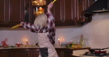 Sarışın, eskrim önlüklü genç bir kadın evde mutfak kaşığıyla merdane takıyor. Neşeli beyaz kadın yemek yapmaktan zevk alıyor. Yaşam tarzı ve neşe. Ciema 4k ProRes Merkezi.