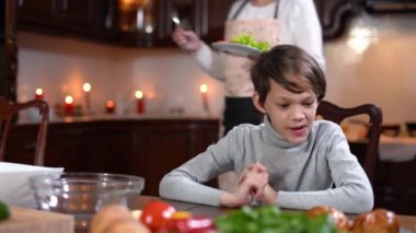 Sağlıklı sebze ve yeşillik getiren tanınmayan bir anne olarak mutfakta oturmuş yemeği bekleyen bir çocuk. Evinde sağlıklı yemek tabağıyla üzgün beyaz bir çocuğun portresi..