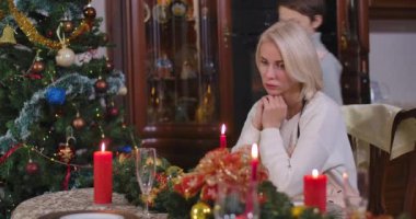 Evdeki süslü Noel masasında oturan üzgün kadını neşelendiren tatlı beyaz çocuk. Yeni yılda annesine evde destek olan olumlu bir oğul. Sinema 4k ProRes Merkezi.