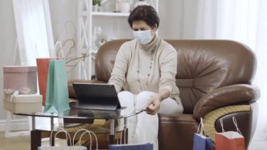 Ödeme kartıyla evde oturan kendinden emin orta yaşlı beyaz alışverişçinin portresi. Covid-19 yüz maskeli esmer kadın koronavirüs hapsinde internetten satın alıyor..