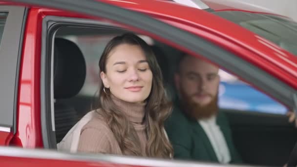 Jonge brunette Kaukasische vrouw met bruine ogen gesloten autodeur en praten met wazige man zittend op de bestuurdersstoel. Portret van een positief koppel kiezen voor een nieuw voertuig in de auto-dealer. Rijkdom concept — Stockvideo