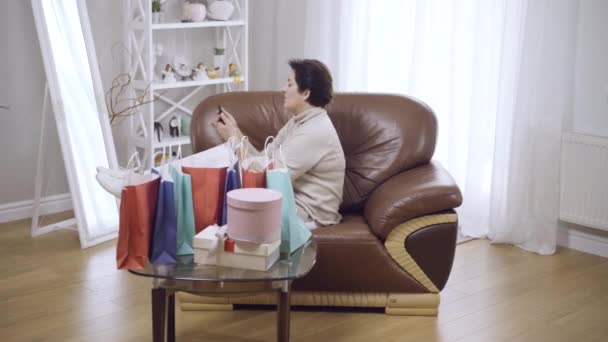 Weite Aufnahme einer entspannten Frau mittleren Alters, die zu Hause auf einer Couch ruht, mit Einkaufstaschen und Schachteln auf einem Glastisch. Unbekümmerte kaukasische Shopperin auf dem Sofa liegend und lächelnd. Lifestyle-Konzept. — Stockvideo