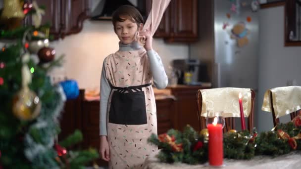 Garçon satisfait positif mettre la main sur les hanches et regarder autour de la maison décorée la veille du Nouvel An. Portrait d'enfant caucasien joyeux dans le tablier se préparant pour la célébration de Noël à l'intérieur. — Video
