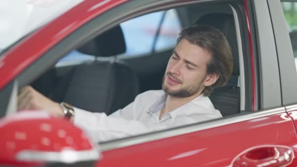 Excited successful young man cheering purchase of new automobile sitting on drivers seat in luxurious car in showroom. Happy Caucasian businessman buying vehicle in dealership. — Stock Video