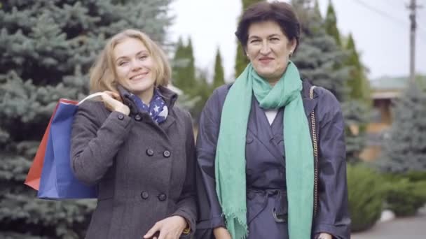 Dvě sebevědomé elegantní ženy pózující s nákupními taškami venku. Portrét krásné štíhlé usmívající se kavkazské mladé a středního věku nakupující při pohledu na fotoaparát chvástání Černý pátek nákupy. — Stock video