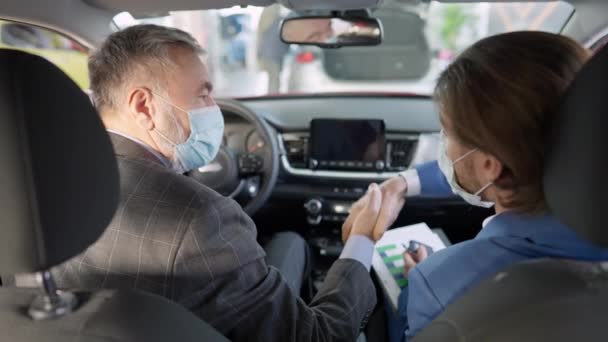 Vista posteriore di uomo anziano fiducioso e giovane rivenditore in maschere facciali Covid-19 che stringono la mano e passano le chiavi seduti all'interno del veicolo. Uomo d'affari l'acquisto di automobili in concessionaria auto su pandemia. — Video Stock