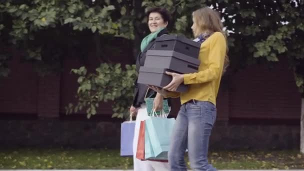 Boční pohled na dvě sebevědomé bělošky s nákupními boxy a taškami, jak si venku povídají. Pozitivní stylové mladé a středního věku dámy vracející se domů na Černý pátek. — Stock video