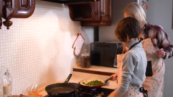 Vedere laterală a fiului caucazian care o ajută pe mama să gătească în bucătărie. Bruneta baiat shaking tigaie la domiciliu indoors. Conceptul culinar și de ajutor . — Videoclip de stoc