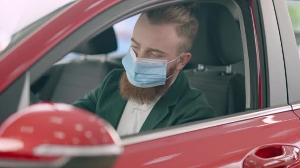 Уверенный молодой бородатый бизнесмен в маске Ковид-19 сидит на водительском сидении в красной машине, осматривая автомобиль. Портрет успешного богатого кавказца, выбирающего автомобиль в автосалоне. — стоковое видео