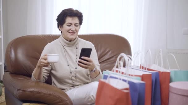 Portret van een zorgeloze stijlvolle vrouw die thuis thee of koffie drinkt na het online winkelen en sms 'en op de smartphone. Positieve glimlach Kaukasische dame zit op de bank surfen Internet. — Stockvideo