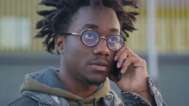 Şehir caddesinde açık havada konuşan yakışıklı Afro-Amerikan bir adamın vesikalığı. Rastalı gözlüklü özgüvenli bir adamın yakın plan portresi. Modern yaşam tarzı.