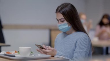 Restoranda akıllı telefon kullanan Covid maskeli güzel bir kadının yan görüntüsü. Kafede yemek yiyen ve internette yavaş çekimde sörf yapan Ortadoğulu muhteşem kadın..