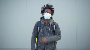 Gözlüklü ve siyah maskeli Afro-Amerikan bir adam başparmağıyla kameraya bakıyor. Şehir merkezindeki gri duvarda duran kendinden emin genç kuryenin portresi..