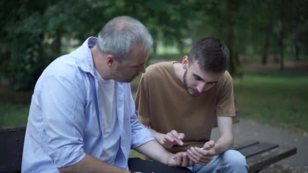 Selbstbewusster junger gutaussehender Mann, der seinen Vater mittleren Alters im Sommerpark über soziale Medien oder Online-Nachrichten unterrichtet. Porträt eines fürsorglichen kaukasischen Sohnes, der Eltern im Freien mit dem Smartphone unterstützt. — Stockvideo