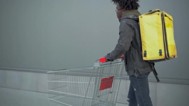 Mladý africký kurýr se žlutou taškou na jídlo tlačí nákupní vozík v městském městě. Muž s dredy přichází, aby si objednal jídlo s sebou. Práce a životní styl. — Stock video
