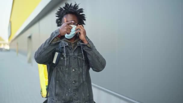 Sebevědomý Afroameričan v Covid-19 s obličejovou maskou mluví po telefonu a chodí s izolovanou taškou na jídlo. Portrét profesionálního kurýra doručujícího objednávku hledajícího klienta v městském městě. — Stock video