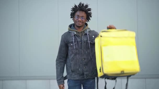 Pemuda Afrika-Amerika yang ceria menunjukkan jempol mengangkat tas pengiriman makanan terisolasi kuning melihat ke kamera. Potret tersenyum kurir berpose di luar kota kota. Konsep layanan. — Stok Video