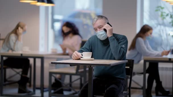 Starší muž v koronavirové masce mluví po telefonu, sedí v restauraci s rozmazanými lidmi v pozadí. Portrét mužského bělocha v kavárně na pandemii Covid-19. Nový normální. — Stock video