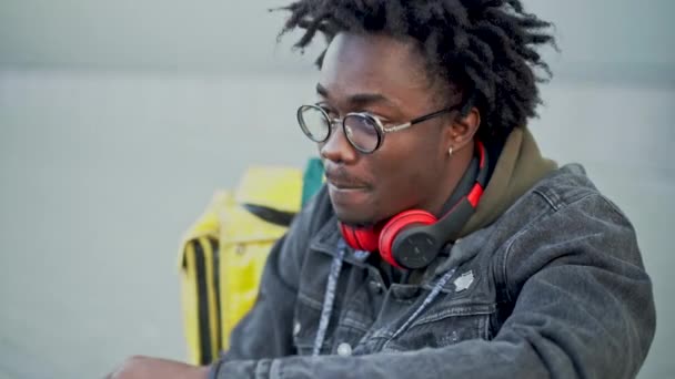 Side view portrait of relaxed African American courier having lunch on urban city street with blurred yellow insulated food delivery bag at background. Job and delivering concept. — Stock Video